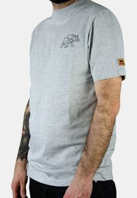 Camiseta de algodón gris de manga corta, con un gráfico de oso geométrico negro en el pecho y una etiqueta de tela color marrón claro en la manga.