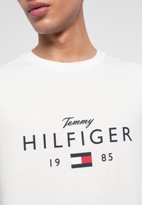Detail zblízka na osobu v bílém tričku s potiskem textu "Tommy Hilfiger" a vlajkovým logem v černé, červené a bílé barvě, včetně čísel 19 a 85.