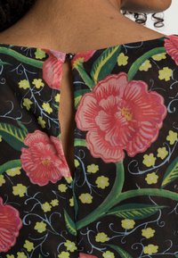 Haut en tissu à motif floral avec de grandes fleurs roses, des feuilles vertes et des accents jaunes sur fond noir ; il dispose d'une fermeture à bouton dans le dos.