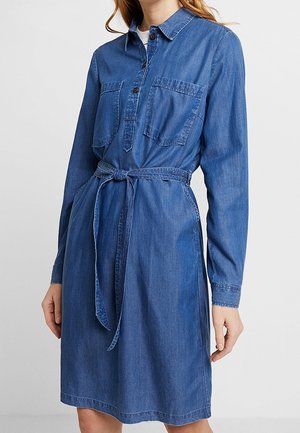 Vrouw draagt een denim shirtjurk met lange mouwen, voorzakken en een gestrikte tailleceintuur, staand tegen een effen lichte achtergrond.