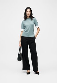 Mujer de pie con bolso negro, vestida con una blusa de manga corta color verde azulado con puños blancos, pantalones anchos negros y zapatos puntiagudos negros.