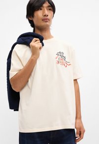 Κρεμ διακοσμημένο βαμβακερό μπλουζάκι με κοντά μανίκια, που διαθέτει πολύχρωμο κεντητό λογότυπο. Άτομο κρατάει μια ναυτική ζακέτα στους ώμους του.