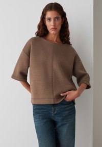 Sweatshirt marron oversize avec manches courtes, tissu texturé, col rond et détails de couture, associé à un pantalon en jean bleu.