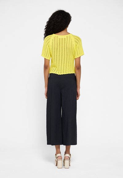 Top corto de punto amarillo con bajo en forma de concha, combinado con pantalones de pierna ancha negros y tacones gruesos beige.