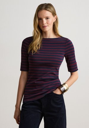 Vrouw met lang blond haar die een marineblauw en bordeaux gestreept shirt met ellebooglange mouwen en donkere broek draagt, staand met één hand in de zak.