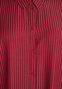 Červená košeľa na gombíky s vertikálnymi čiernymi pruhmi, klasickým golierom a textúrovanou tkaninou. Päť červených gombíkov je zarovnaných nadol po prednej strane.