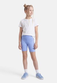 Κορίτσι με πλεγμένα μαλλιά, φορώντας λευκό μπλουζάκι, ανοιχτό μπλε σορτς ποδηλασίας Puma, λευκές κάλτσες και ασορτί ανοιχτό μπλε αθλητικά παπούτσια, στέκεται μπροστά σε λευκό φόντο.