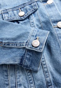 Manica di giacca di denim azzurro chiaro con cuciture visibili, bottone metallico inciso con testo del marchio e parte della patta della tasca anteriore visibile.