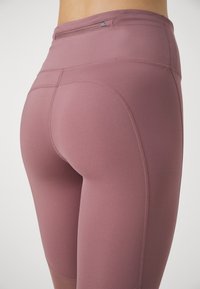 Close-up van een persoon die hooggerokte mauve legging draagt met achterste stiksels en een klein ritszakje in de tailleband.