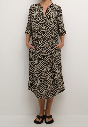 Femme portant une robe fluide beige jusqu'aux genoux avec un imprimé zèbre noir, manches courtes, poches latérales, et sandales noires sans lacets.