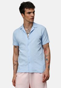 Camisa de manga corta texturizada en azul claro con cuello y cierre de botones. Lleva un estilo casual sobre pantalones cortos rosa claro.