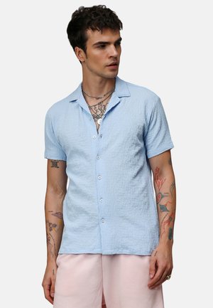 Camisa de manga corta texturizada en azul claro con cuello y cierre de botones. Lleva un estilo casual sobre pantalones cortos rosa claro.