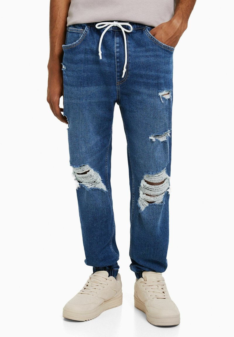 Distressed blue denim jogger pants with white drawstring, featuring multiple torn details, and cuffed ankle design, paired with light beige shoes.