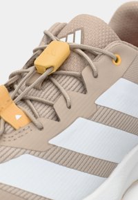 Μπεζ αθλητικό παπούτσι με πλέγμα ανώτερου μέρους, λευκές ρίγες και κίτρινες μεταλλικές θηλιές για τα κορδόνια. Διαθέτει υφή στην επιφάνεια και επένδυση στον γιακά.