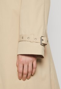 Beige trenchcoat ermer som har en spenne- og øyeskruedetalj. Glatt stoff, tilpasset passform, synlig hånd som hviler på ermet.