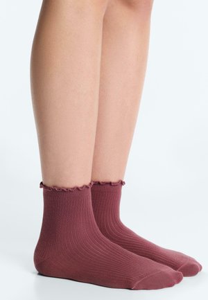 Pair of feet wearing ribbed, ankle-length dusty rose socks with ruffled cuffs against a plain light background.