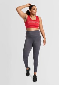 Röd kort sportbh med "HUMMEL"-logotyp, kombinerad med högmidjade grå leggings och svarta träningsskor; mjukt tyg, åtsittande design.