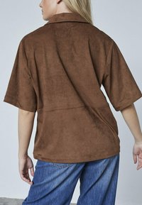 Kvinde iført løs brun ruskinds kortærmet skjorte og blå jeans, vist fra ryggen mod en ensfarvet lys baggrund.