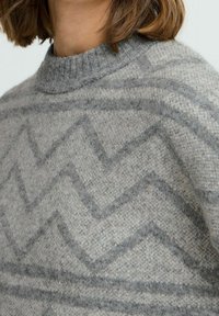Femme portant un pull gris en tricot avec un col montant et un motif subtil en zigzag dans des tons de gris plus foncés.