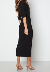 Zwarte, geribbelde, midi-jurk met puffmouwen, getailleerde pasvorm en details in de zijnaad. Gemaakt met doorzichtige sandalen met hakken.