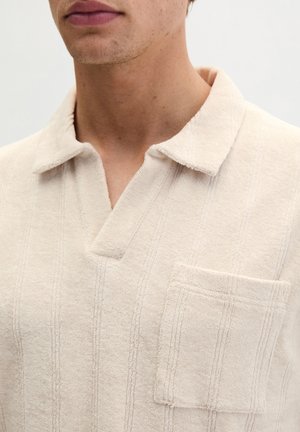 Beiges strukturiertes Kurzarmhemd mit offenem Kragen und Brusttasche, getragen von einer Person, die vom Hals bis zur oberen Brust gezeigt wird.