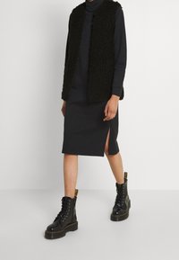 Czarna kamizelka z futra sztucznego noszona na czarnej sukience z długim rękawem i bocznym rozcięciem, zestawiona z czarnymi botkami za kostkę z wiązaniem i grubą podeszwą.