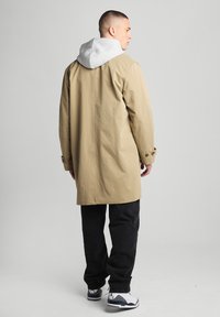 Beige waterproof coat with a gray hood, featuring a straight cut, buttoned cuffs, and black pants, viewed from the back.