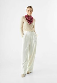 Beige trui, witte high-waisted geplooide broek en een roze geweven sjaal geknoopt om de nek. Zachte texturen en neutrale tinten.