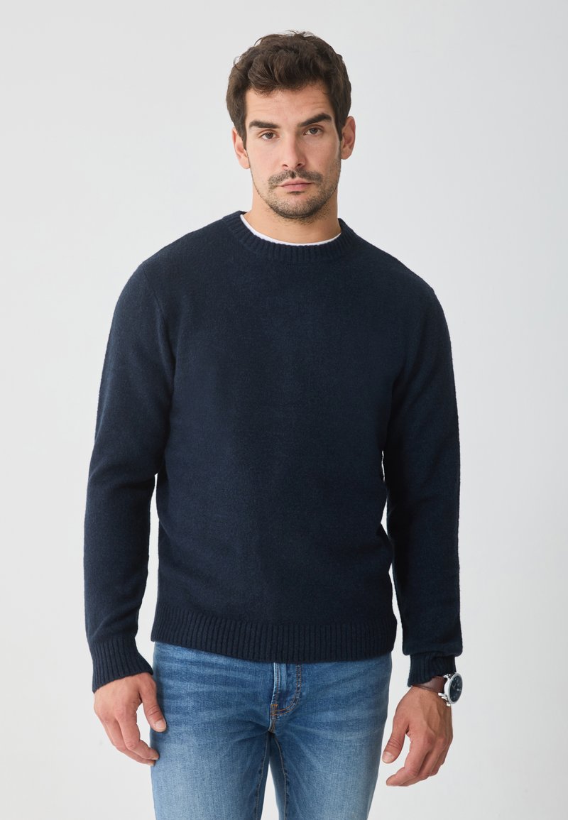 Marineblauer Strickpullover mit rundem Halsausschnitt sowie gerippten Bündchen und Saum, kombiniert mit hellblauen Jeans. Weiche Textur, schlichtes Design.
