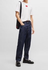 Hvid t-shirt med et rødt logo, mørkeblå vide jeans, sorte sko og en sort skuldertaske med røde detaljer. Enkel og afslappet stil.