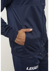Chaqueta atlética azul marino hecha de tela suave, con cierre de cremallera y bolsillos laterales con un patrón texturizado. Logo "LEGEA" visible.