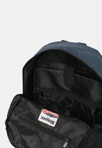 Zaino nero con parte superiore blu navy, caratterizzato da un compartimento principale con zip, tasche interne e un'etichetta in tessuto. Materiale resistente con una texture liscia.