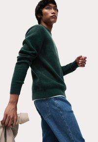 Grüner Strickpullover mit gerippter Textur, kombiniert mit blauen Jeans. Das Model hält eine beige Jacke und steht leicht schräg.