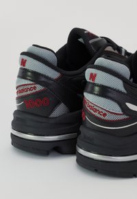 Paire de baskets New Balance 1000 noires et grises avec des détails de logo rouges et des talons épais rembourrés sur une surface blanche.