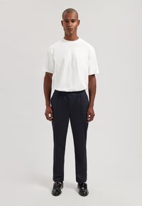 Hvit bomulls-T-skjorte og mørke bukser med folder. Buksene har elastisk midjebånd og glatt tekstur. Sorte sko fullfører antrekket.