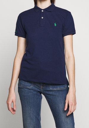 Polo bleu marine avec col à revers, trois boutons, logo vert brodé et manches courtes. Porté avec un jean bleu.