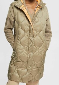 Pikowany płaszcz do kolan w oliwkowym zielonym kolorze, z pomarańczową podszewką i kapturem, bocznymi kieszeniami oraz zapięciem na guziki.