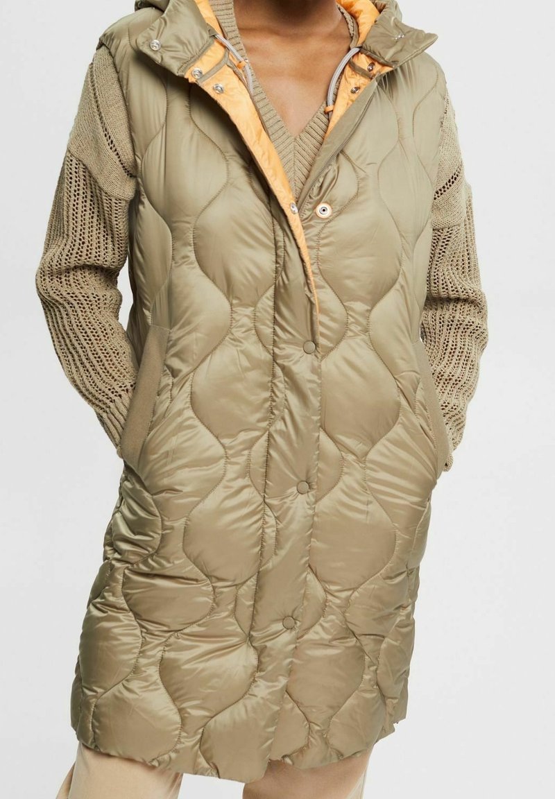 Pikowany płaszcz do kolan w oliwkowym zielonym kolorze, z pomarańczową podszewką i kapturem, bocznymi kieszeniami oraz zapięciem na guziki.