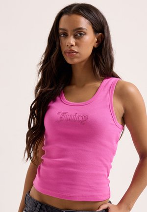 Frau mit langem dunklem Haar, trägt ein eng anliegendes pinkfarbenes Tanktop mit dem Schriftzug „Juicy“ in stilisierter Schrift auf der Vorderseite, posiert vor einem einfarbigen Hintergrund.