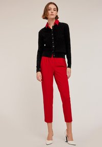 Czarny żebrowany kardigan z czerwonym kołnierzem i guzikami, w połączeniu z czerwonymi dopasowanymi spodniami i białymi szpilkami z ostro zakończonym noskiem, zaprezentowany na modelce.