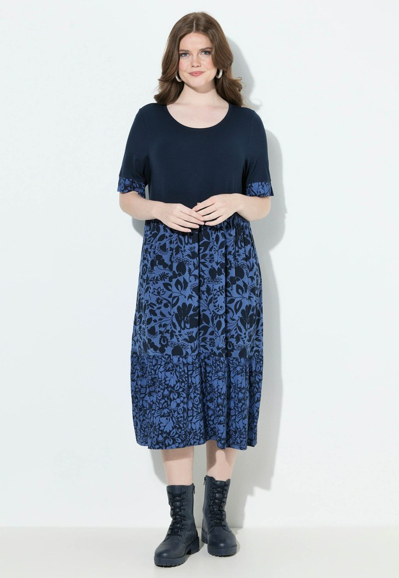 Granatowa sukienka w kwiaty z krótkimi rękawami, o wielowarstwowym kroju. Połączona z czarnymi, sznurowanymi botkami. Materiał wydaje się miękki i lekki.