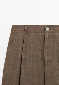 Pantalons en lin marron avec une finition texturée, dotés d'une fermeture à boutons, de plis à l'avant et de poches latérales.
