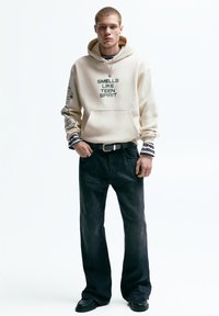 Beige hoodie met de tekst "SMELLS LIKE TEEN SPIRIT", zwarte jeans en zwarte schoenen. De hoodie heeft grafische accenten op de mouwen en de voortas.