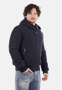 Giacca a vento blu navy con cappuccio realizzata in tessuto testurizzato, dotata di chiusura con zip frontale, tasche laterali e polsini a costine. Indossata con jeans azzurri chiari.