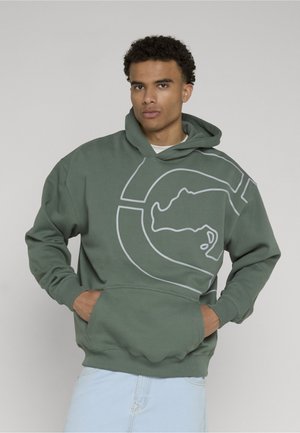 Homme aux cheveux bouclés portant un sweat à capuche vert avec une grande silhouette abstraite d'animal, mains dans la poche avant, jean bleu clair.
