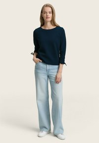 Donna in piedi con una mano nella tasca dei pantaloni a palazzo azzurri chiari, indossa una maglietta a maniche lunghe blu navy e sneakers bianche su uno sfondo semplice.