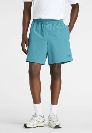 Homme portant un short de sport turquoise, des chaussettes blanches, des baskets blanches et une chemise blanche, debout avec une main dans la poche.