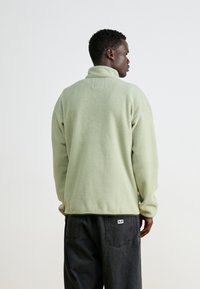 Grønn fleecejakke med høy krage, myk tekstur og avslappet passform. Kombinert med mørke jeans med detalj på baklommen.