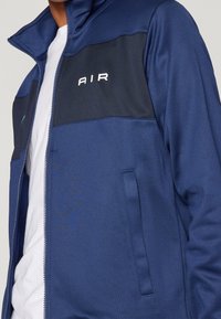 Marinblå zip-up jacka med svarta detaljer, med ståkrage, sidofickor och ordet "AIR" i vitt på bröstet. Slät tygkvalitet.