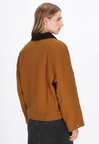 Bruine gewatteerde jas met een zwarte corduroy kraag, met een relaxte pasvorm en knoopaccenten bij de mouwen. De textuur lijkt glad.